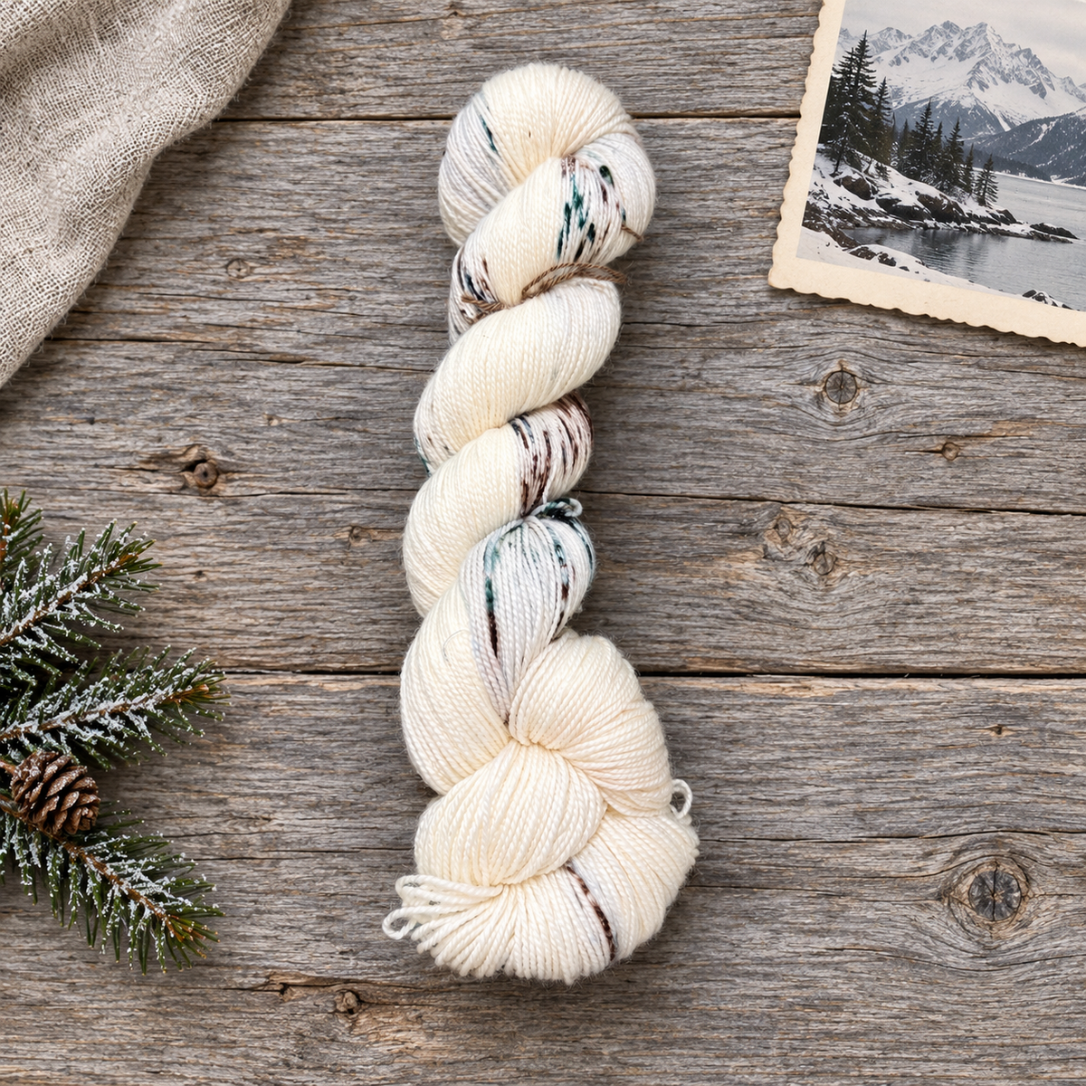 The Great Alone cream hand-dyed yarn with sparse green and brown markings on gray wood beside frosted evergreen and an Alaskan winter landscape print.