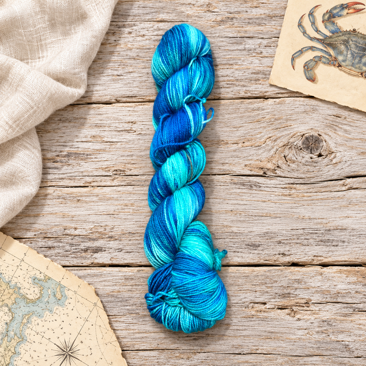 Chesapeake hand-dyed yarn skein in blue, turquoise, and aqua on weathered wood beside a vintage crab illustration and nautical chart.