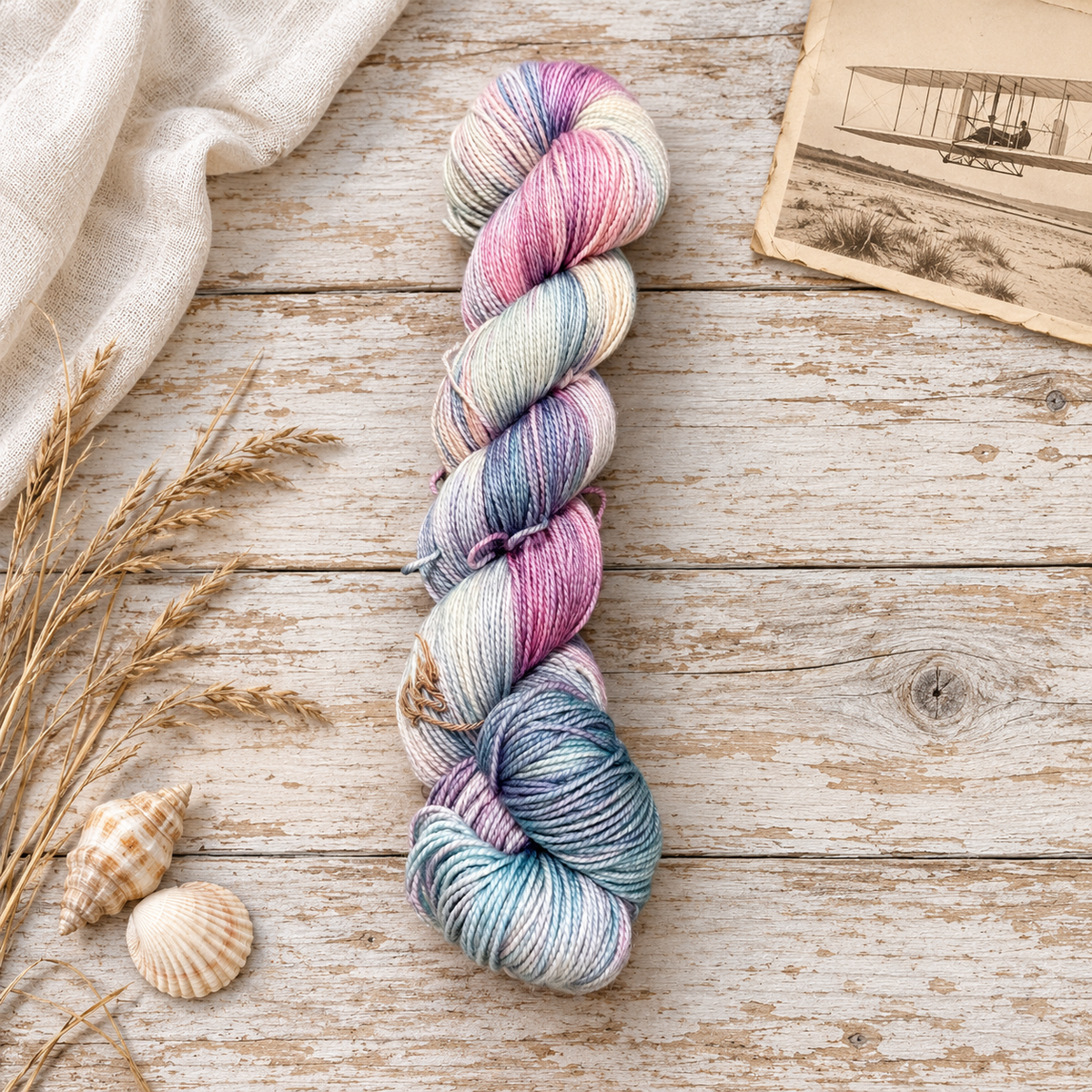 Kitty Hawk hand-dyed yarn in pink, mauve, cream, and blue-gray on weathered wood with seashells, dune grasses, and a vintage aviation print.
