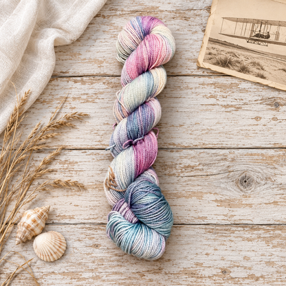Kitty Hawk hand-dyed yarn in pink, mauve, cream, and blue-gray on weathered wood with seashells, dune grasses, and a vintage aviation print.
